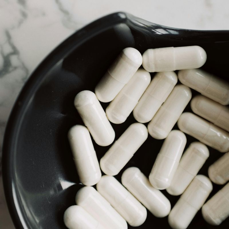 Capsules blanches de complément alimentaire versées dans une assiette noire sur fond de marbre blanc