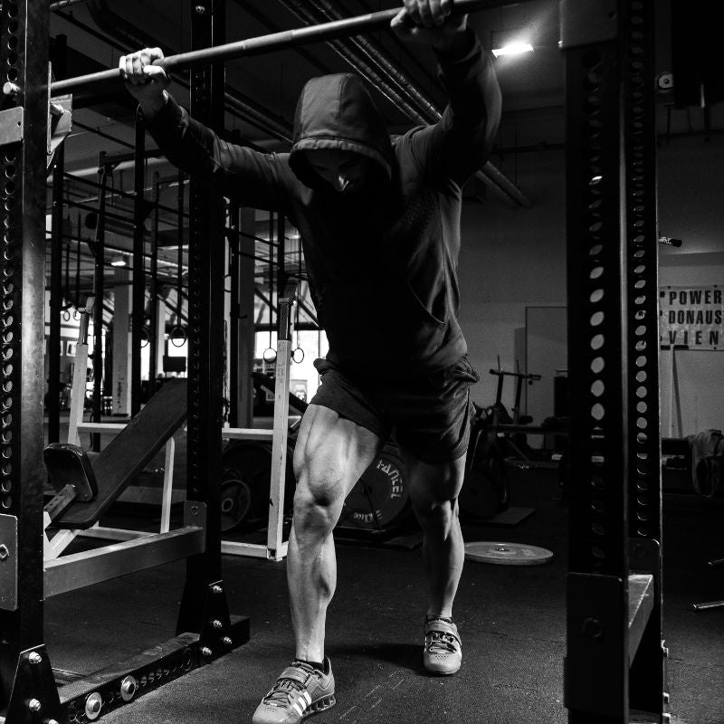 Homme musclé en hoodie poussant une barre lourde sur rack de squat dans une salle de musculation en noir et blanc