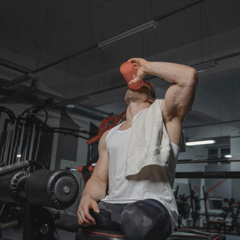 Homme musclé en salle de gym buvant une boisson de récupération dans un shaker rouge, essuiefront blanc, après entraînement