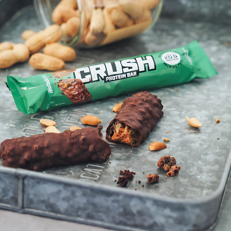Barre protéinée CRUSH BioTechUSA emballage vert avec barre coupée en deux sur plateau métallique, entourée de cacahuètes, indiquant 20 g de protéines sans huile de palme