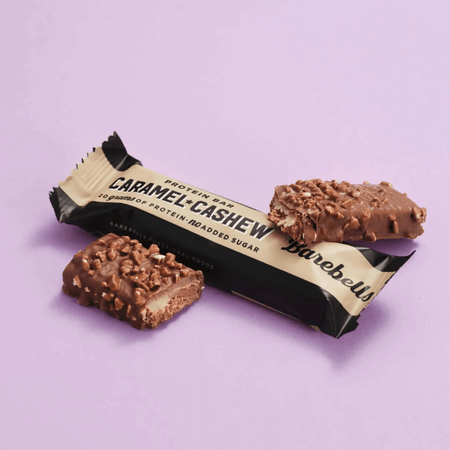 Barre protéinée Barebells caramel noix de cajou, recouverte de chocolat au lait et d’éclats croustillants, posée sur un fond rose avec emballage beige et noir mentionnant 20 g de protéines sans sucres ajoutés
