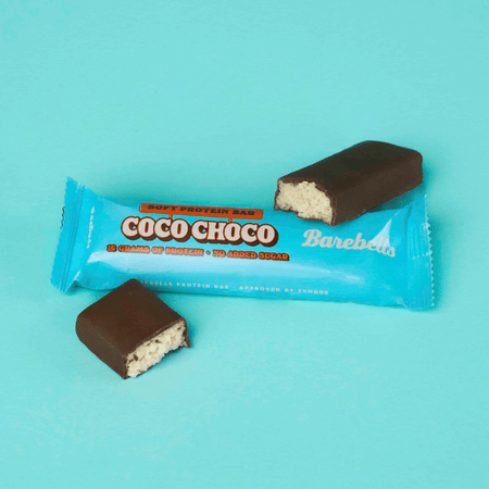 Barre protéinée Barebells Coco Choco à la texture moelleuse, enrobée de chocolat noir avec intérieur coco, présentée en morceaux sur un fond turquoise avec emballage bleu indiquant 16 g de protéines et sans sucres ajoutés