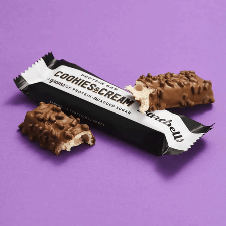 Barre protéinée Barebells cookies and cream, enrobée de chocolat au lait avec morceaux croquants, présentée coupée sur un fond violet avec emballage noir et blanc indiquant 20 g de protéines sans sucres ajoutés