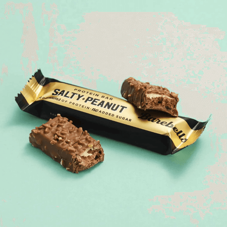 Barre protéinée Barebells salty peanut, chocolat au lait parsemé de morceaux de cacahuètes, posée sur un fond vert avec emballage doré et noir marqué 20 g de protéines sans sucres ajoutés
