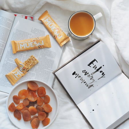 Scène petit-déjeuner cosy avec barres Protein Muesli BiotechUSA abricot, tasse de thé, abricots secs et carnet ouvert portant l’inscription Enjoy every moment, le tout posé sur du linge blanc