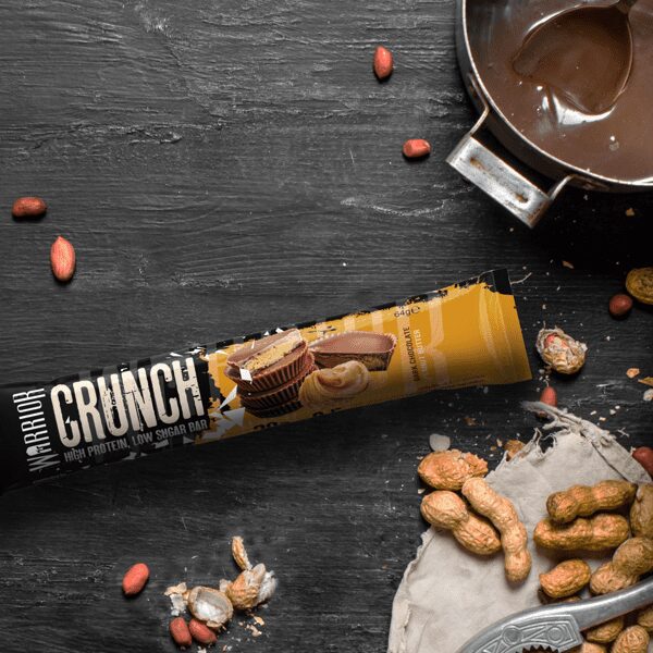 Barre protéinée Warrior Crunch saveur peanut butter disposée sur planche noire avec cacahuètes entières et sauce chocolat dans une casserole