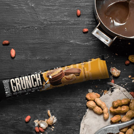 Barre protéinée Warrior Crunch saveur peanut butter disposée sur planche noire avec cacahuètes entières et sauce chocolat dans une casserole