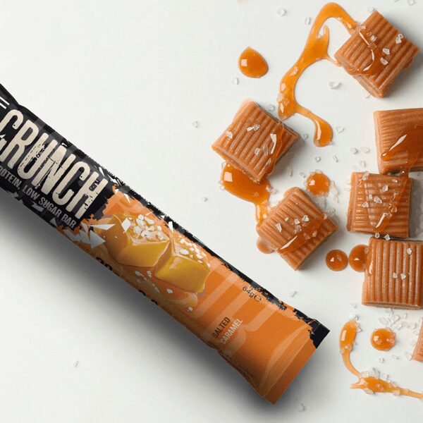 Barre protéinée Warrior Crunch Salted Caramel sur fond blanc accompagnée de carrés de caramel nappés de caramel liquide et parsemés de cristaux de sel