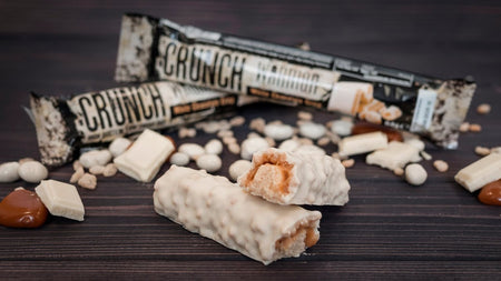 Barres protéinées Warrior Crunch enrobées de chocolat blanc avec une barre coupée en deux laissant apparaître le cœur caramel, entourées de morceaux de chocolat blanc et de billes croustillantes sur fond bois sombre