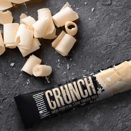 Barre protéinée Warrior Crunch chocolat blanc entourée de copeaux de chocolat blanc sur surface pierre foncée