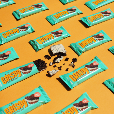 Disposition à plat de multiples barres BioTechUSA Crispy Cocoa sur fond orange avec une barre coupée en morceaux laissant apparaître le cœur croustillant clair