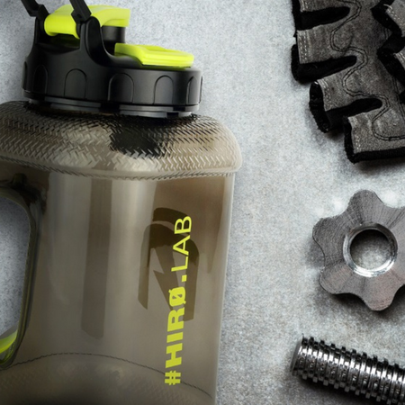 Gros plan du bidon HIRO.LAB gris fumé posé sur un sol de salle de sport à côté de gants et de disques de musculation