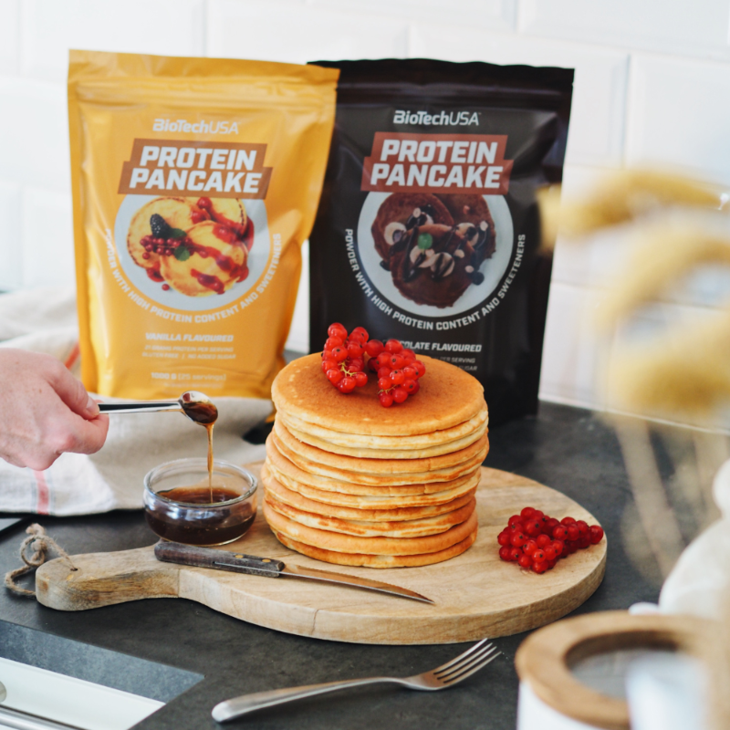 Pile de pancakes dorés sur planche en bois avec sirop et groseilles rouges, devant deux sachets BioTechUSA Protein Pancake vanille et chocolat
