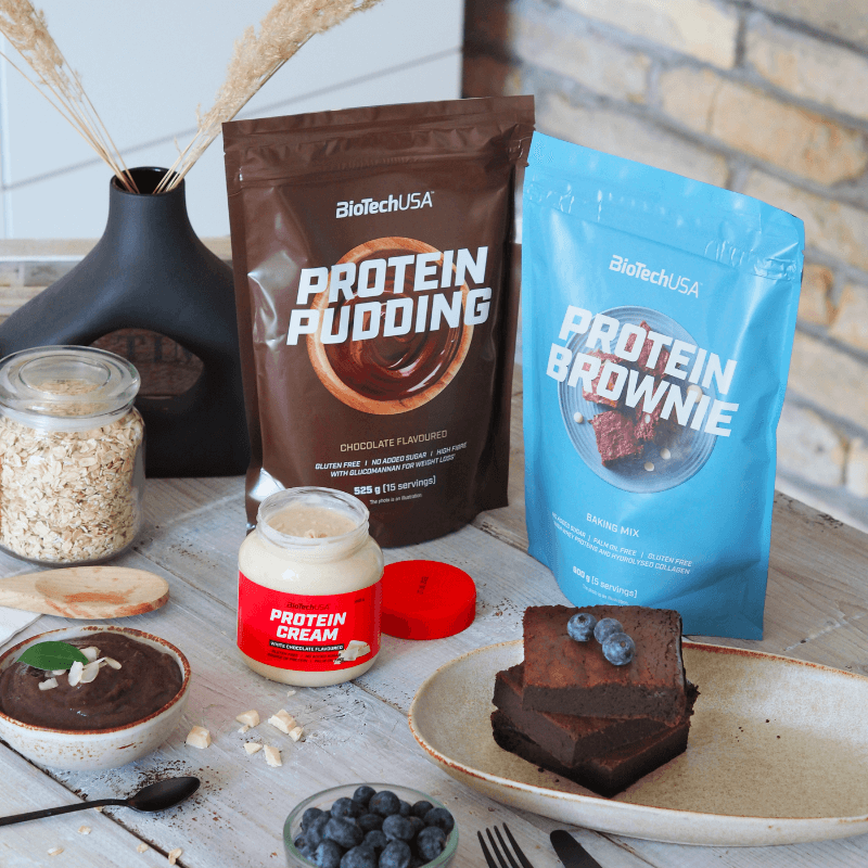 Sachets BiotechUSA Protein Pudding et Protein Brownie posés sur une table avec bols de dessert chocolaté, flocons d’avoine et myrtilles