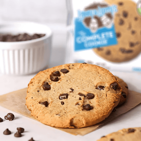 Gros plan d’un cookie Lenny & Larry’s aux pépites de chocolat posé sur un papier cuisson avec des pépites autour et l’emballage flou en arrière-plan