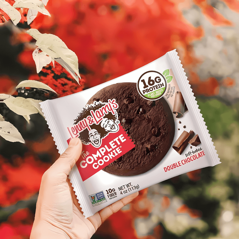 Sachet Lenny & Larry’s The Complete Cookie double chocolat tenu à la main devant un fond automnal rouge, montrant un gros cookie tout chocolat et 16 g de protéines.​
