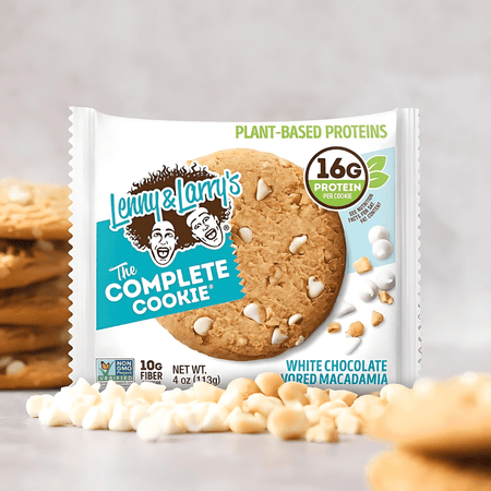 Cookie Lenny & Larry’s The Complete Cookie saveur chocolat blanc et macadamia en sachet blanc et turquoise avec éclats de chocolat blanc et noix de macadamia au premier plan, 16 g de protéines végétales