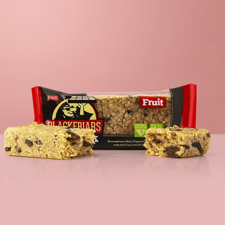 Barre flapjack Blackfriars aux fruits, riche en flocons d’avoine et morceaux de fruits secs, posée devant l’emballage rouge et noir portant la mention suitable for vegans sur fond rose