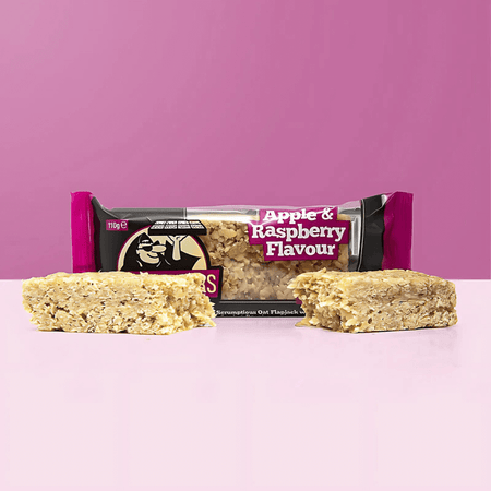 Barre flapjack Blackfriars saveur pomme framboise, texture moelleuse aux flocons d’avoine, posée devant l’emballage fuchsia et noir sur fond rose