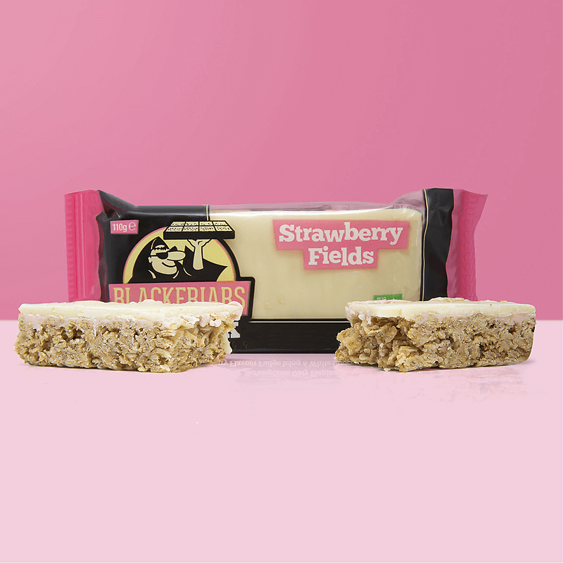 Barre flapjack Blackfriars Strawberry Fields avec nappage blanc, présentée en deux morceaux devant l’emballage rose et noir sur fond rose pastel