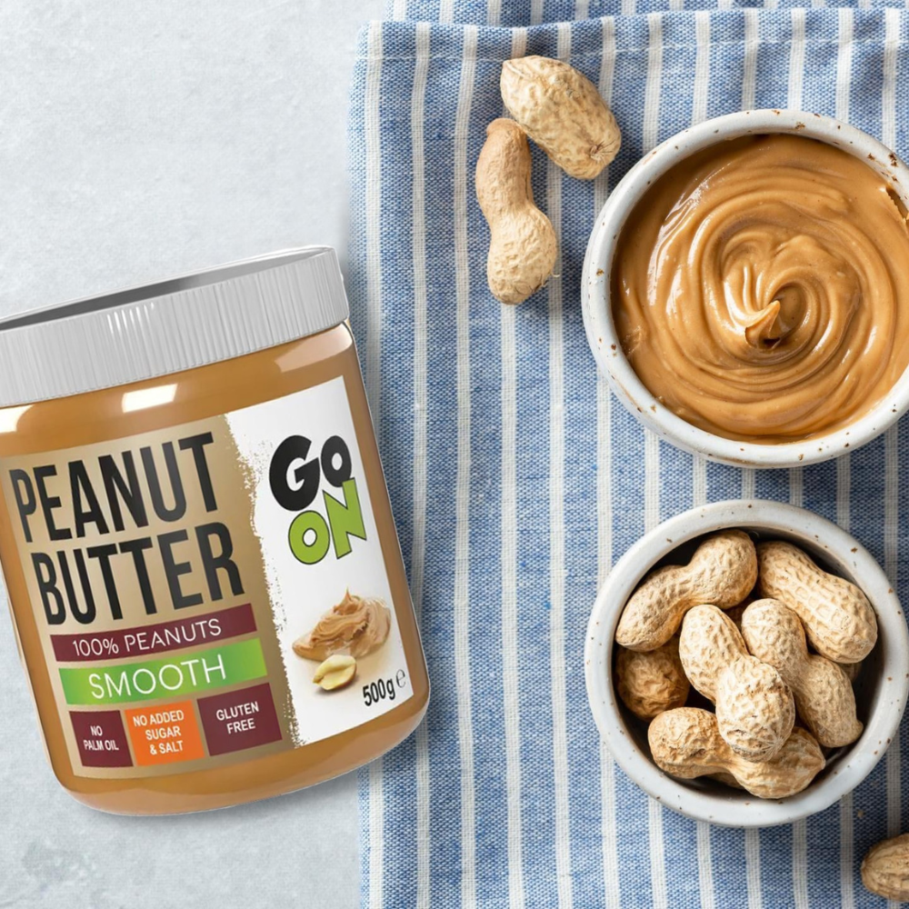 Pot de beurre de cacahuète Go On Smooth 100% cacahuètes posé sur un torchon rayé avec un bol de beurre de cacahuète et des cacahuètes entières