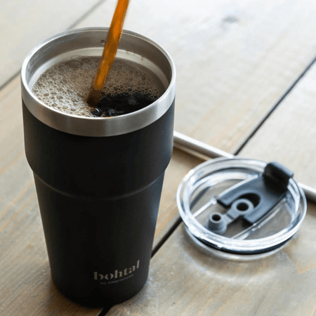 Mug de voyage isotherme Bohtal 600 ml noir rempli de café chaud avec couvercle posé à côté sur une table en bois.

