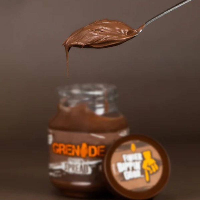 Pot de pâte à tartiner Grenade chocolat au lait ouvert avec couvercle posé devant et cuillère tenant une portion de pâte chocolatée onctueuse au-dessus du pot.