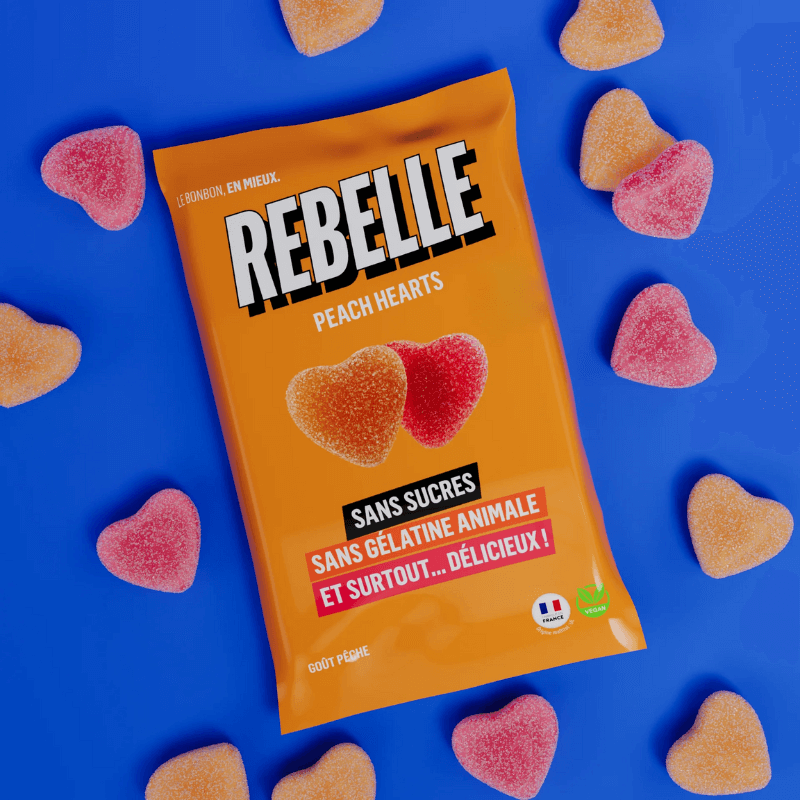 Sachet orange de bonbons Rebelle Peach Hearts sans sucres ni gélatine animale entouré de cœurs gélifiés goût pêche sur fond bleu vif