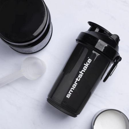 Shaker Smartshake 800 ml noir posé sur une surface blanche à côté d’une cuillère doseuse et d’un pot de poudre