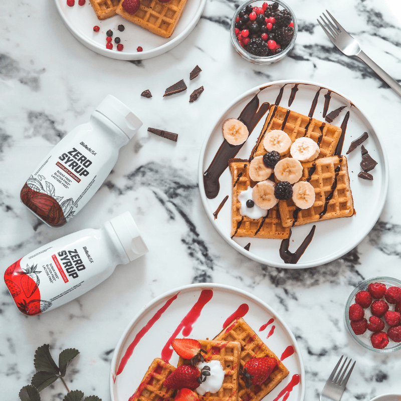 Assiettes de gaufres garnies de fruits rouges et banane nappées de sirop avec deux bouteilles de Zero Syrup BioTechUSA posées sur un plan de travail en marbre