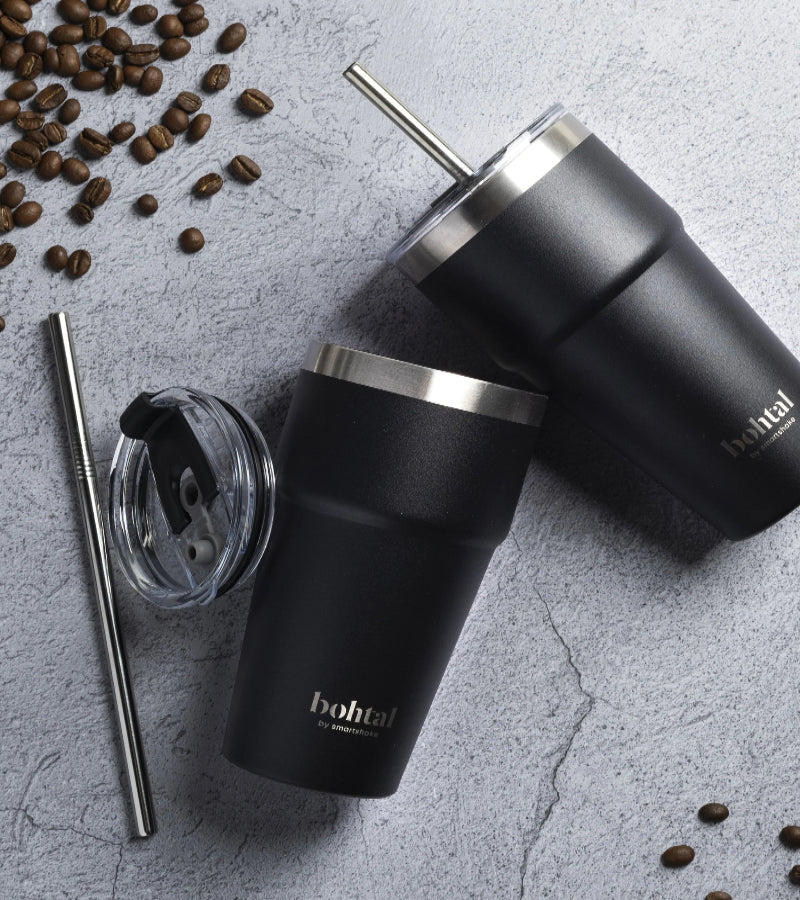Deux tumblers isothermes Bohtal noirs avec paille inox posés sur un plan de travail gris entourés de grains de café et d’un couvercle supplémentaire