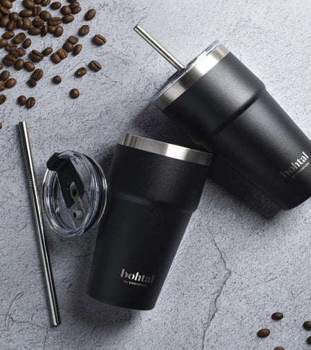Deux tumblers isothermes Bohtal noirs avec paille inox posés sur un plan de travail gris entourés de grains de café et d’un couvercle supplémentaire