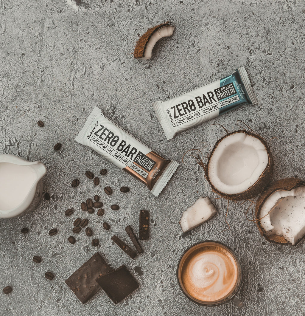 Mise en scène de barres protéinées Zero Bar BioTechUSA avec café, éclats de chocolat et noix de coco sur fond en béton, illustrant des snacks riches en protéines et pauvres en sucre